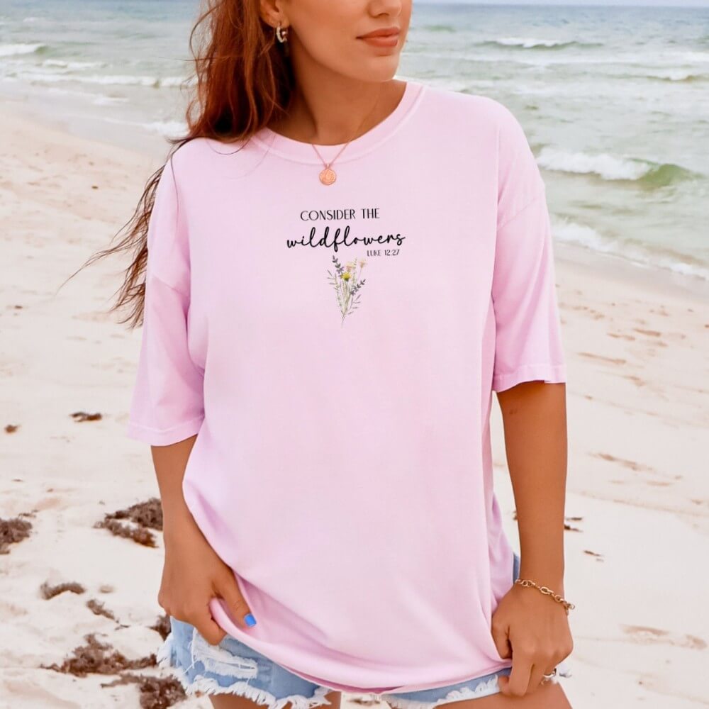 Consider the Wildflowers t-shirt. Short sleeve, crew neck unisex t-shirt (pink color) with a wildflower printed on the front chest and Luke 12:27 verse "Consider The Wildflowers"