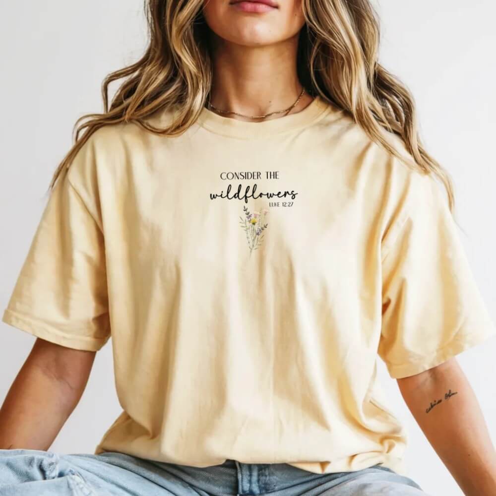 Consider the Wildflowers t-shirt. Short sleeve, crew neck unisex t-shirt (butter color) with a wildflower printed on the front chest and Luke 12:27 verse "Consider The Wildflowers"