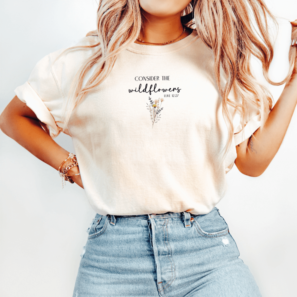 Consider the Wildflowers t-shirt. Short sleeve, crew neck unisex t-shirt (ivory color) with a wildflower printed on the front chest and Luke 12:27 verse "Consider The Wildflowers"