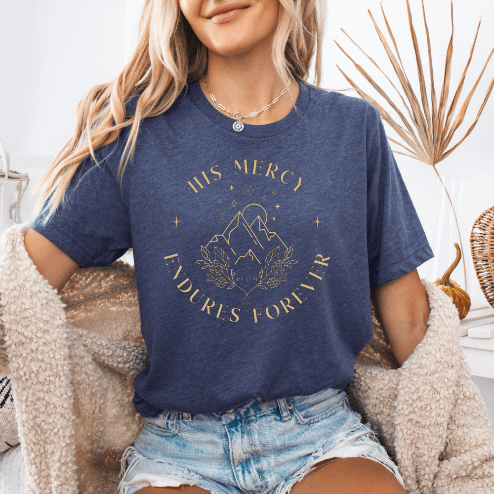His Mercy Endures forever short sleeve women's t-shirt heather midnight navy.  Design of mountains in tan with "His Mercy Endures Forever" printed on the front around the design. "Psalm 136" printed in small font.