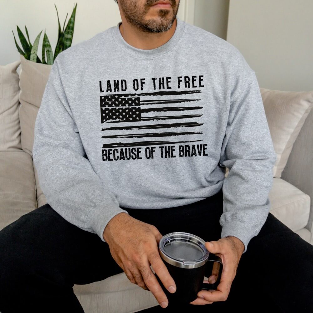 Unisex Land of the Free sweatshirt sport grey.  An American flag is printed on the front in black with "Land of the free because of the brave" above and below the flag.
